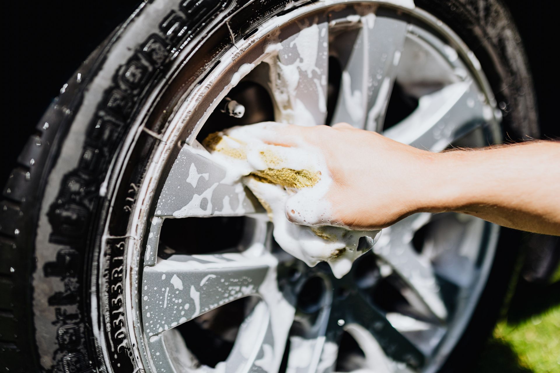A close-up image of a hand scrubbing a car rim and tire with suds, emphasizing car wash detail.
