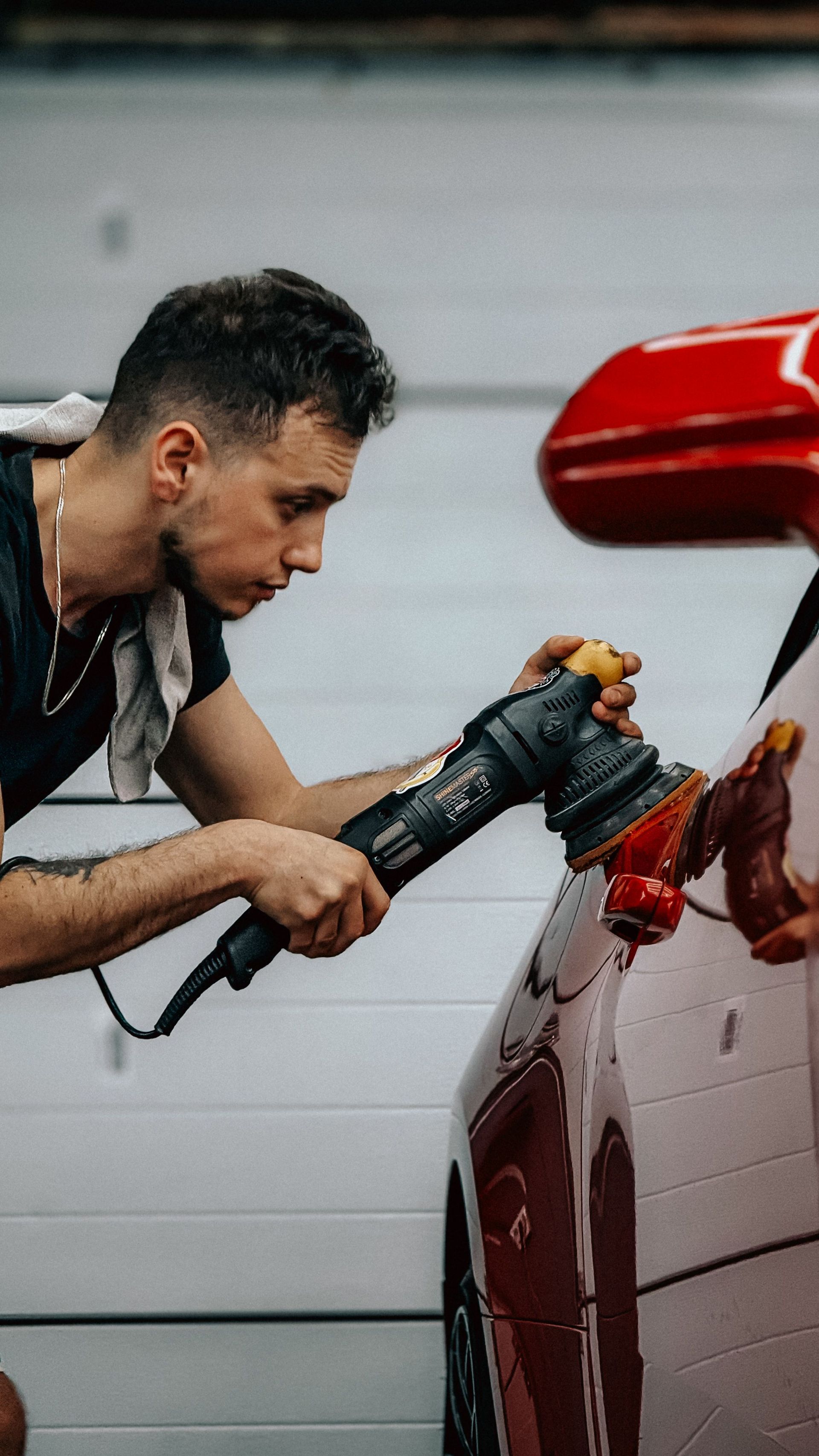 Focused man diligently polishing a red car with a buffer in a garage, highlighting car care.