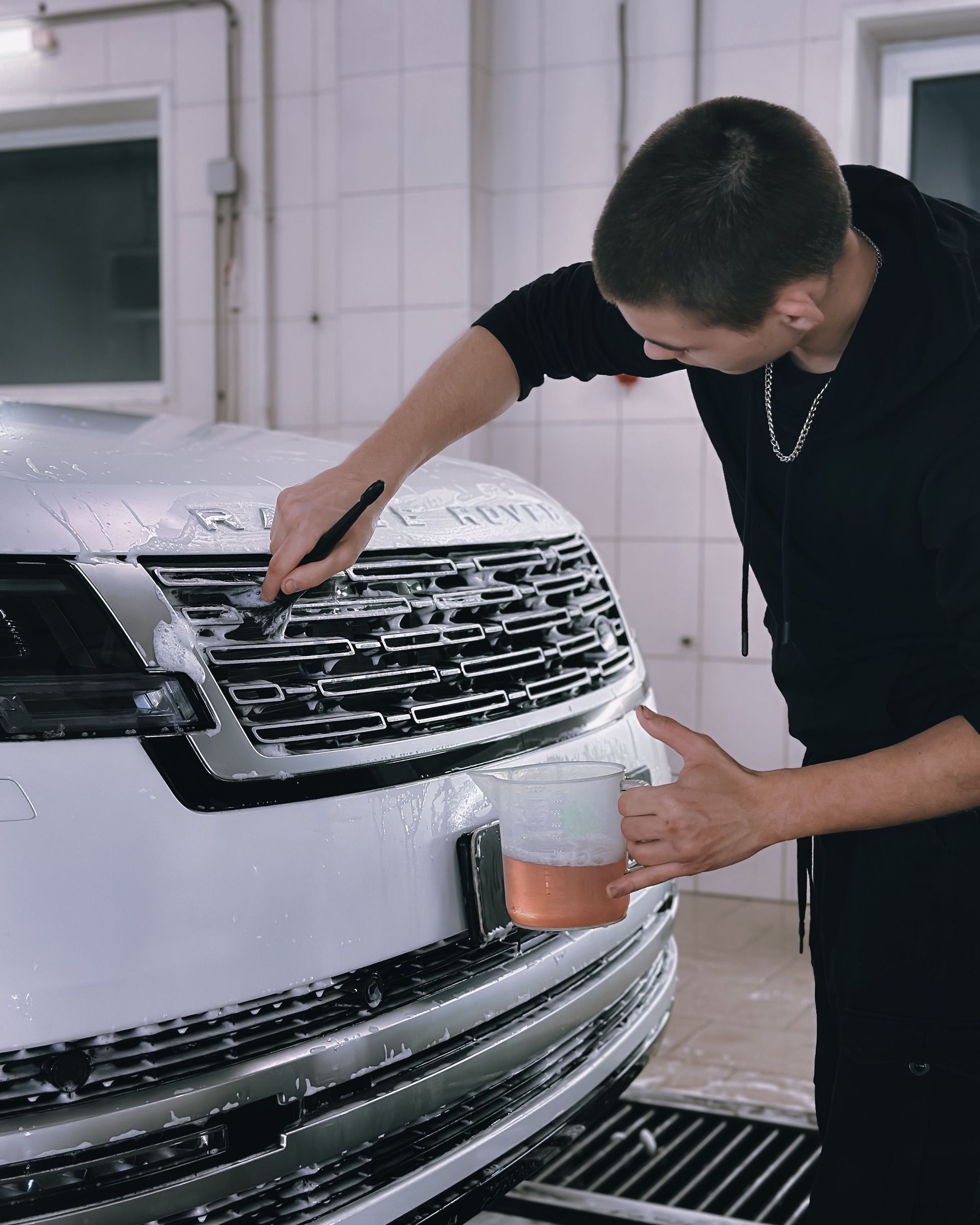 A man washing a white luxury vehicle's grill in an indoor garage, focusing on cleanliness.
